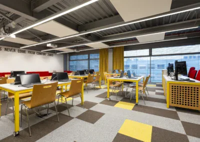 Alternativní text: Učebna s moderními počítači, židlemi a velkými okny s žlutými záclonami ve školícím centru.